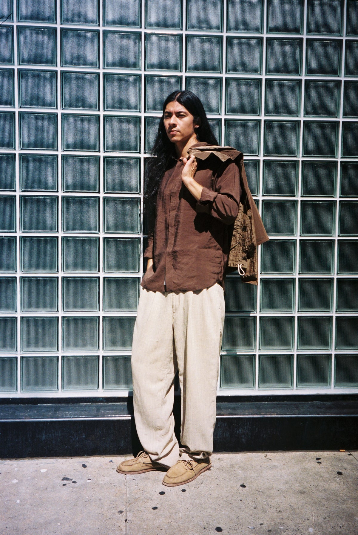 A model in a brown shirt, cream trousers and Colbo x Sperry boat shoes against a glass block wall