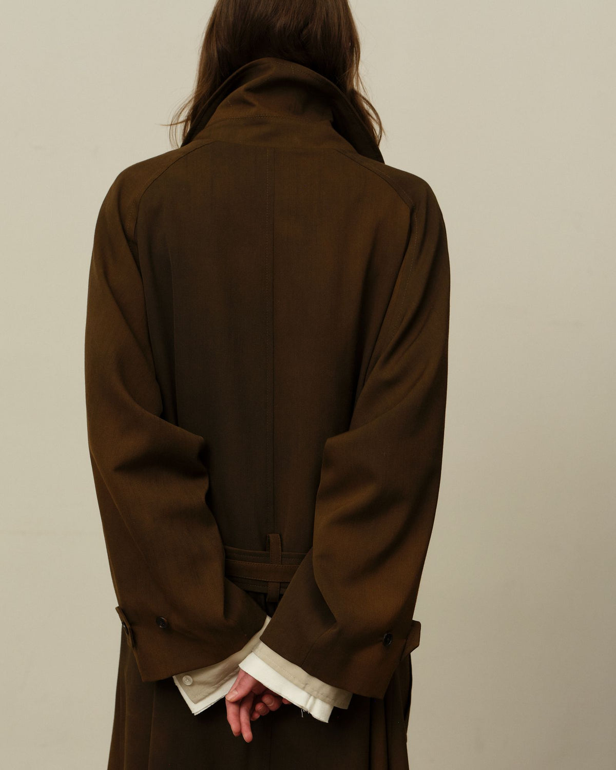 Back view of a model in a long olive brown trench coat, hands clasped behind