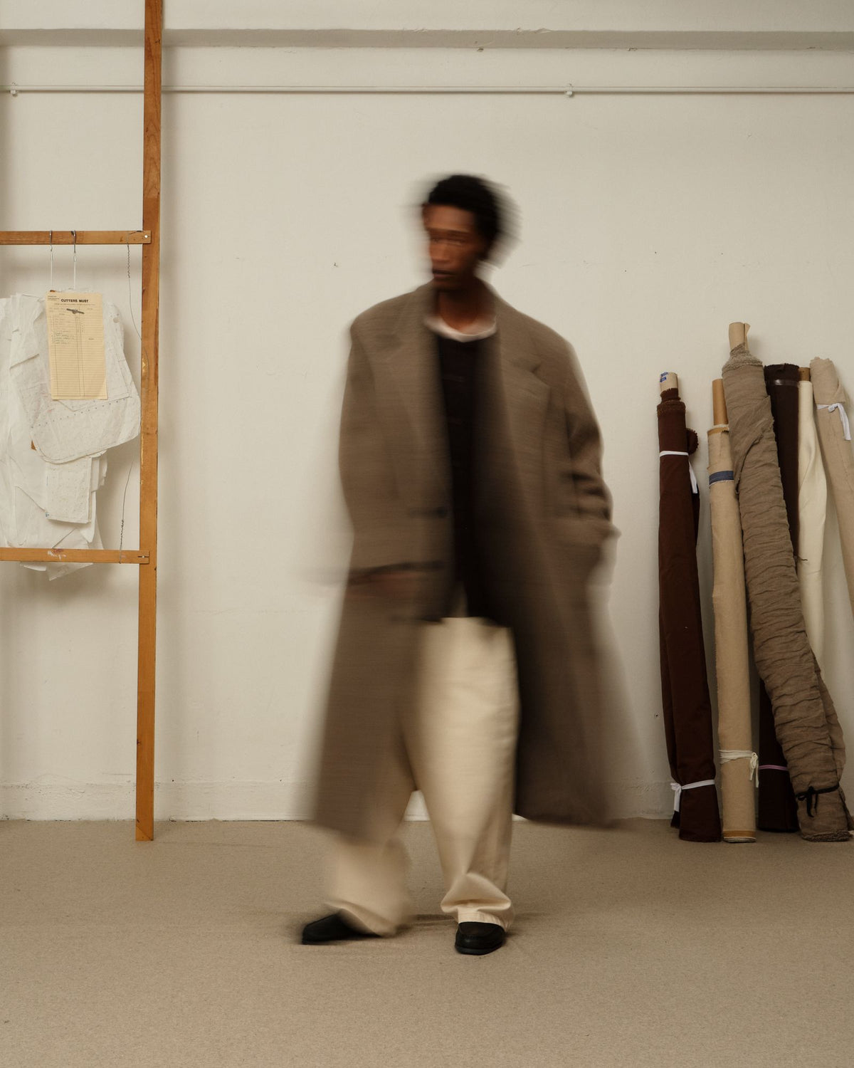 A blurred model walks past in a long taupe coat and cream wide-leg pants