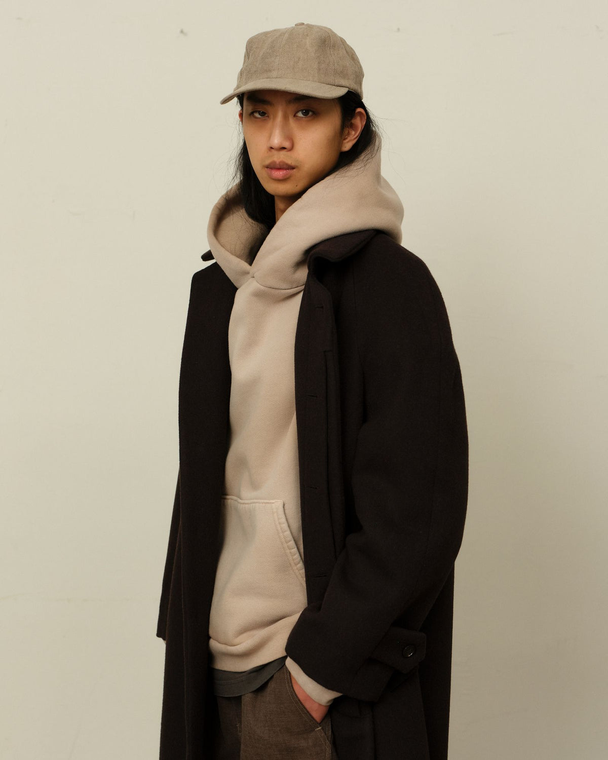 A model in a long dark brown overcoat over an oatmeal hoodie and a greige cap