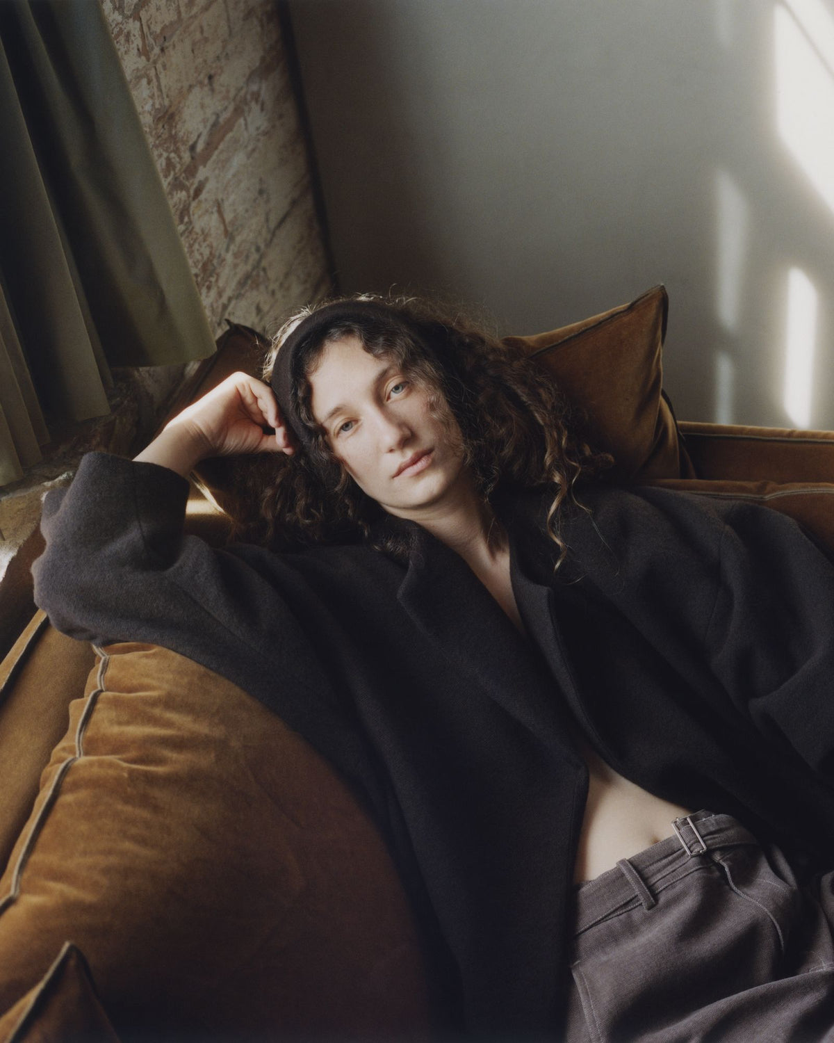 A model resting on an ochre velvet sofa in a dark jacket and Colbo Belt Pants in Brushed Dark Brown Canvas