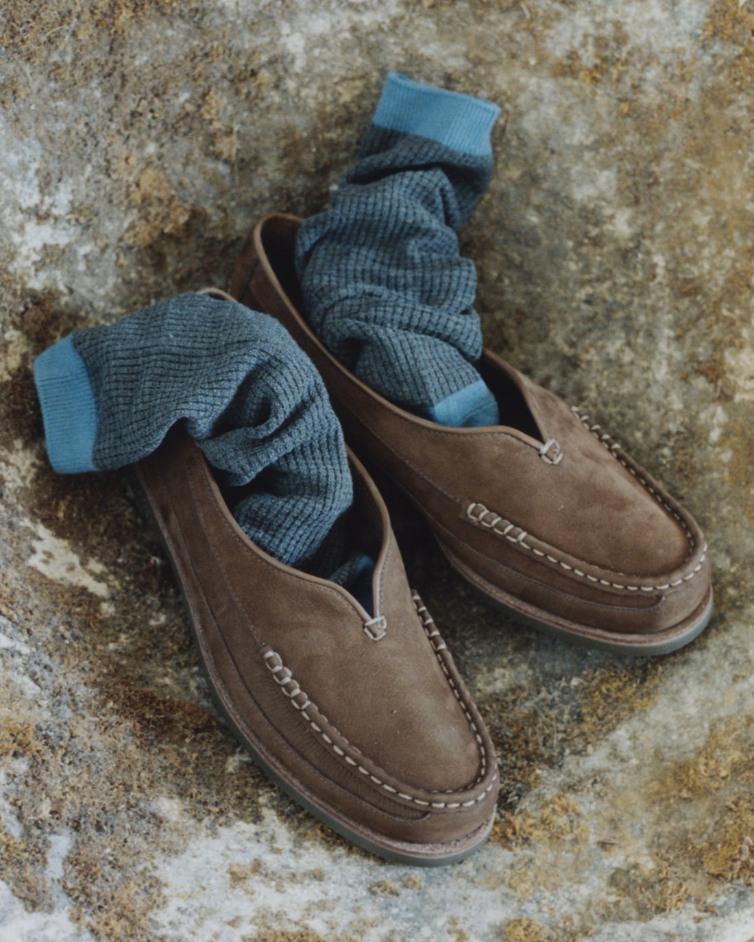 Chocolate nubuck Sirah loafers with blue socks on a stone table