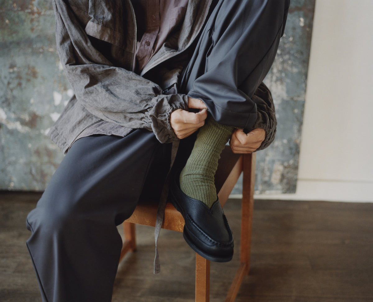 Someone on a wooden chair pulling on a black Sirah loafer over a green sock