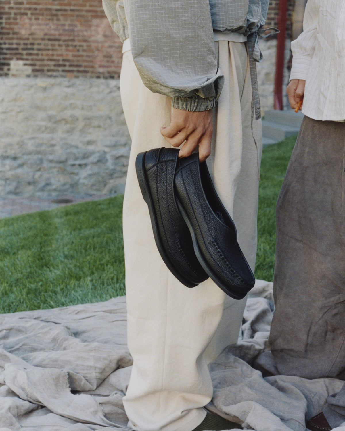 Holding a pair of black Sirah loafers on a lawn by a brick wall