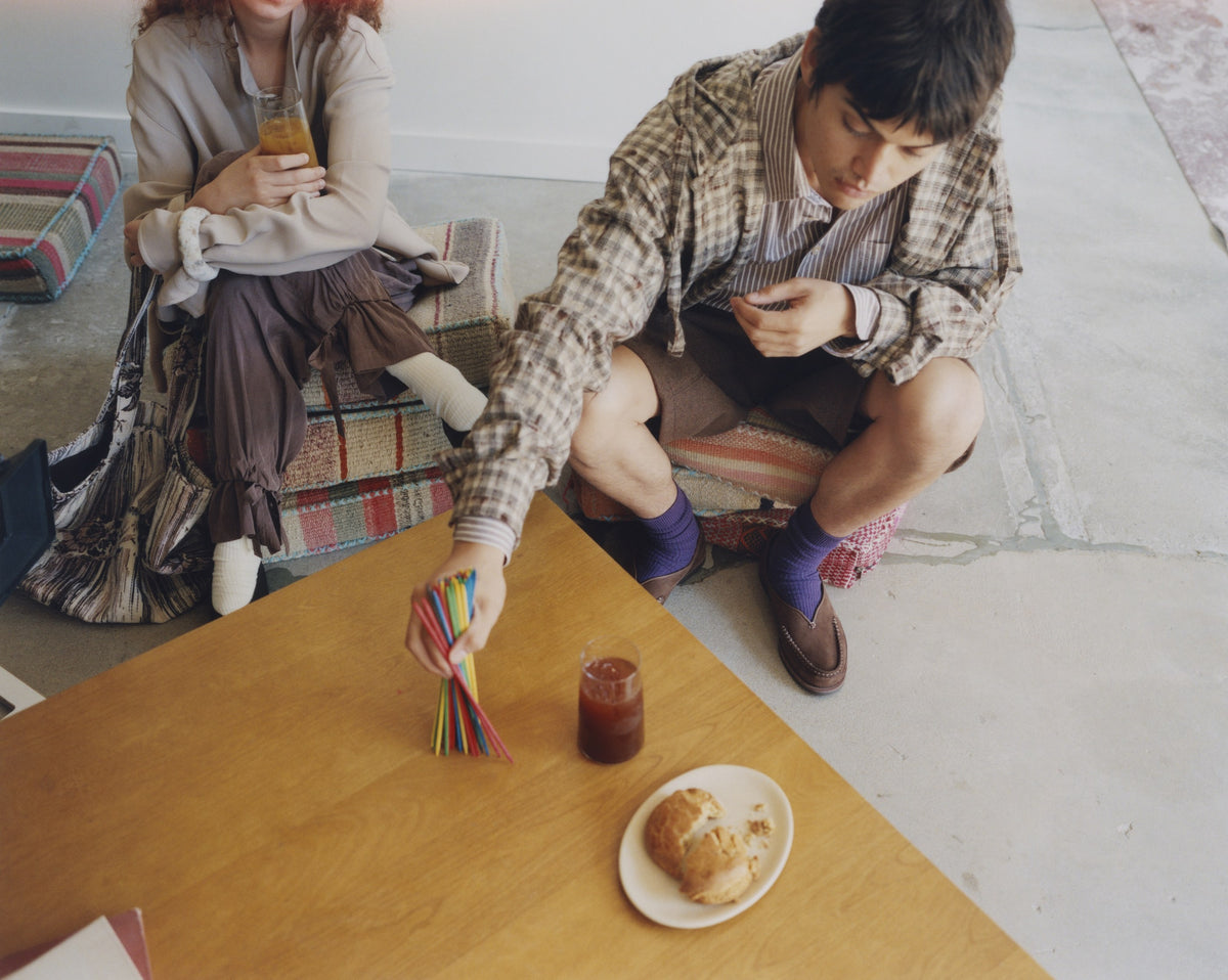 Two friends around a low wooden table, one in chocolate Sirah loafers and purple socks
